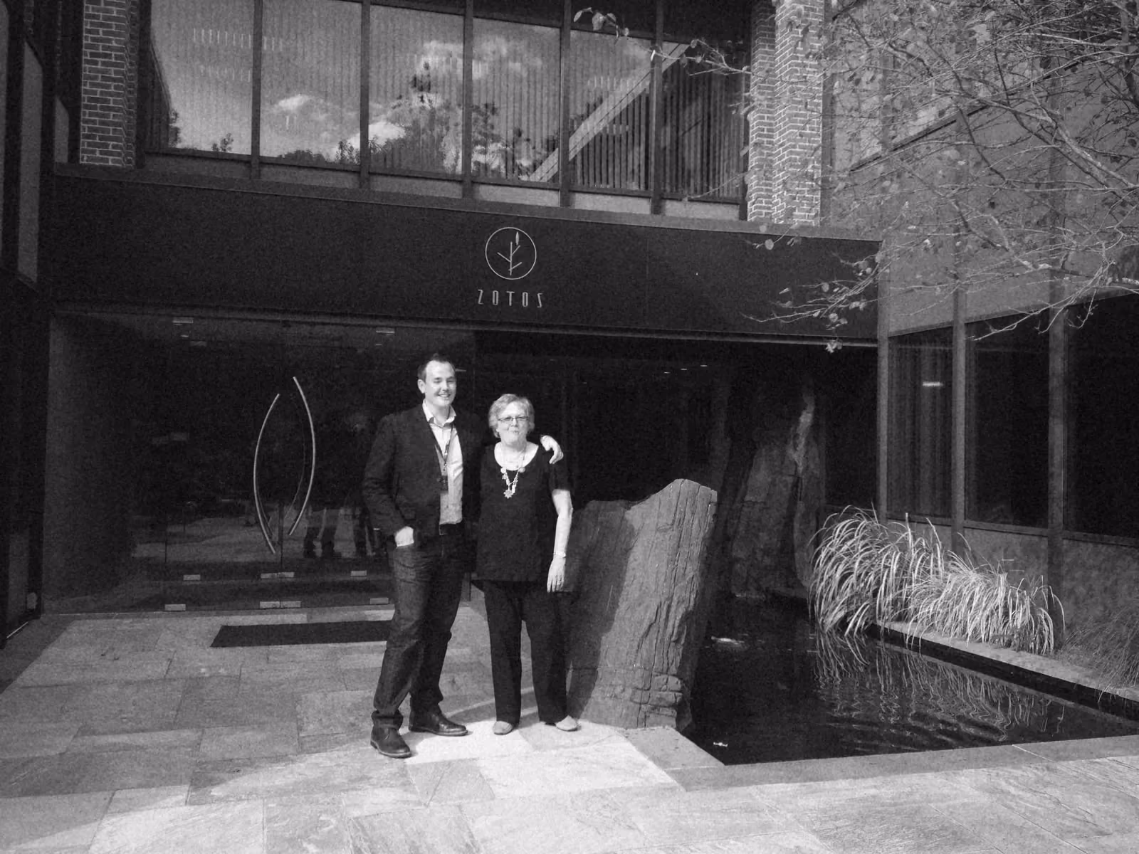 Ben and a colleague standing arm in arm outside the Zotos head office, beside a slate water feature