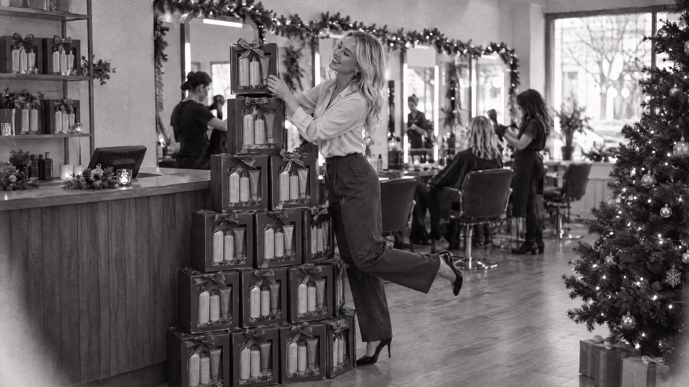 A busy salon at Christmas, a sales rep on tiptoe with one foot kicked up behind her, placing the top pack on a pyramid of boxed haircare Christmas gift packs beside the reception counter