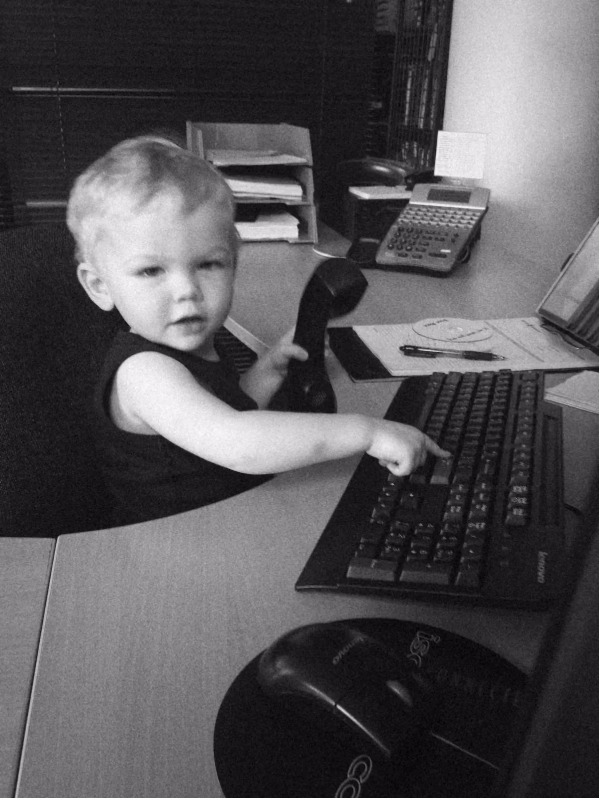 A toddler at an office desk holding a phone handset and pressing a key on the keyboard